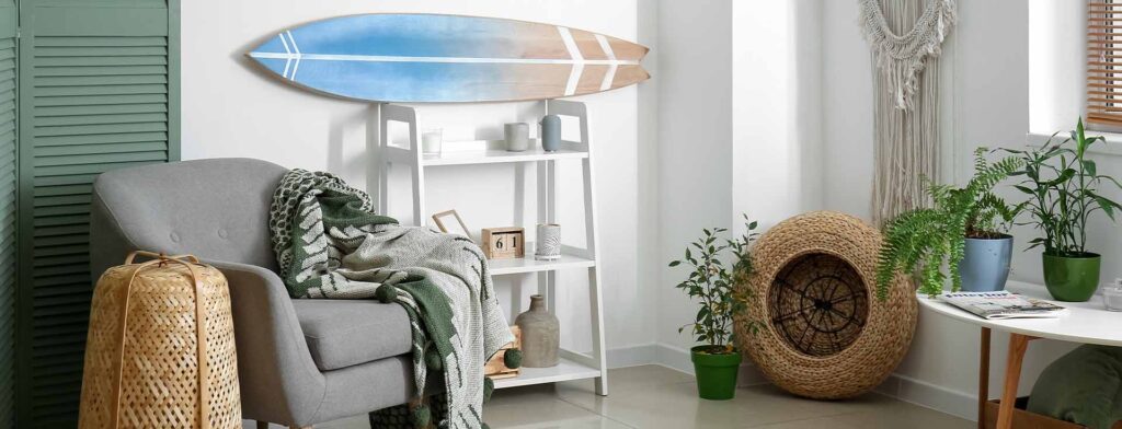 Casa com decoração praiana, há plantas em mesas e no chão, uma prancha de surfe pendurada na parede e uma poltrona com uma manta em cima.
